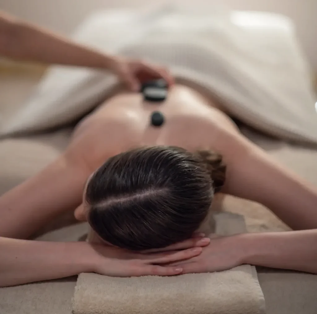 Člověk leží obličejem dolů na masážním stole s ručníkem a nechává si masírovat záda horkými kameny, což podporuje relaxaci a celkovou pohodu.